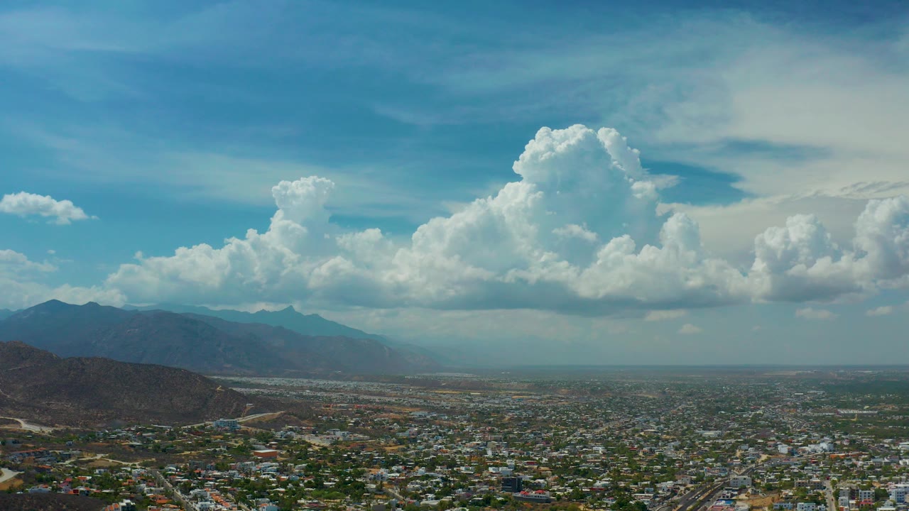 Opening aerial frame over Los Cabos, Mexico, showing dense low-rise city blocks, brown hills, distant mountains, a blue sky, large white clouds, and no visible people.