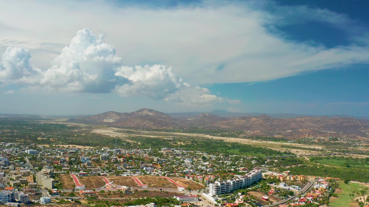 Early aerial frame of Los Cabos with city blocks in the foreground, green scrubland through the center, desert mountains beyond, and no visible people.