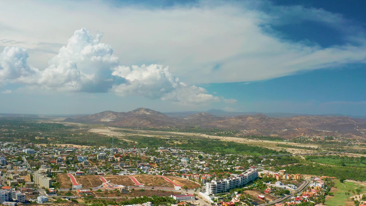 Aerial view across Los Cabos with a city edge at the bottom, rugged hills across the horizon, and a large white cloud mass overhead with no visible people.