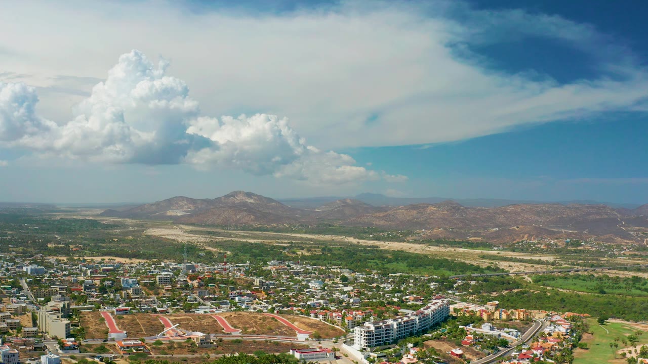 A wide aerial view over Los Cabos, Mexico shows low buildings, roads, green land, distant hills, and large white clouds, with no clearly visible people.