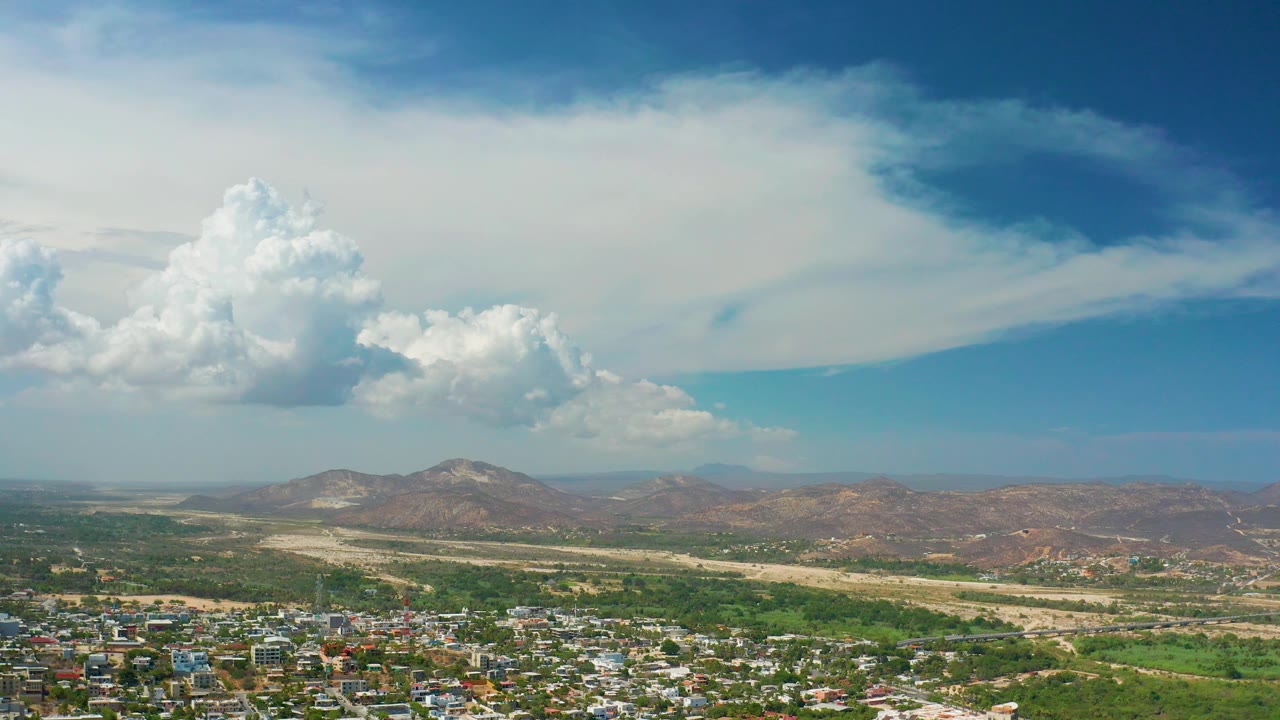Wide aerial landscape with blue sky, white clouds, mountains, trees, and rooftops.