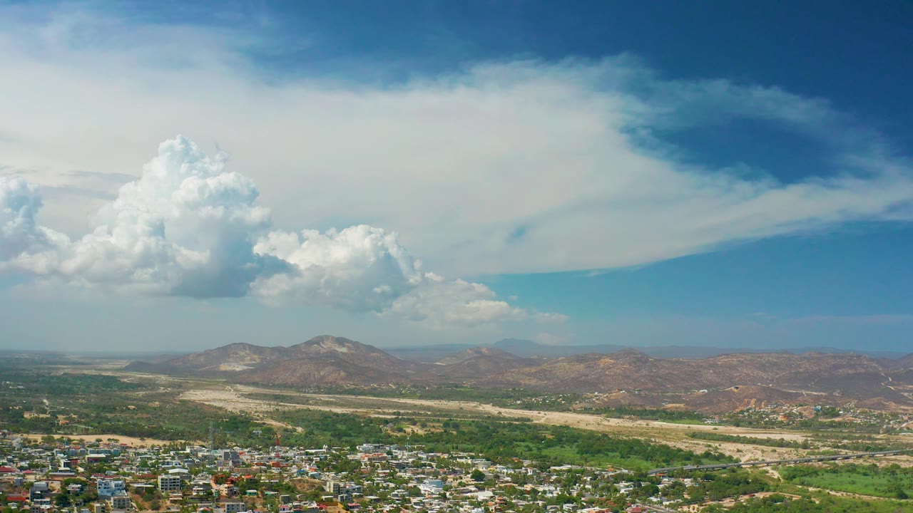 Opening aerial frame over Los Cabos, Mexico, with town buildings along the bottom edge, a green valley and dry wash beyond, brown hills on the horizon, and no visible people.