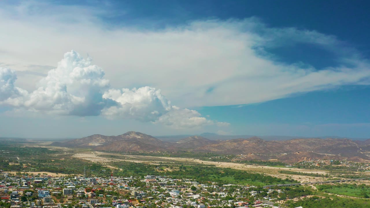 Los Cabos town remains at the lower edge while the valley, light-colored wash, low hills, and a large cloud bank dominate the frame, with no visible people.