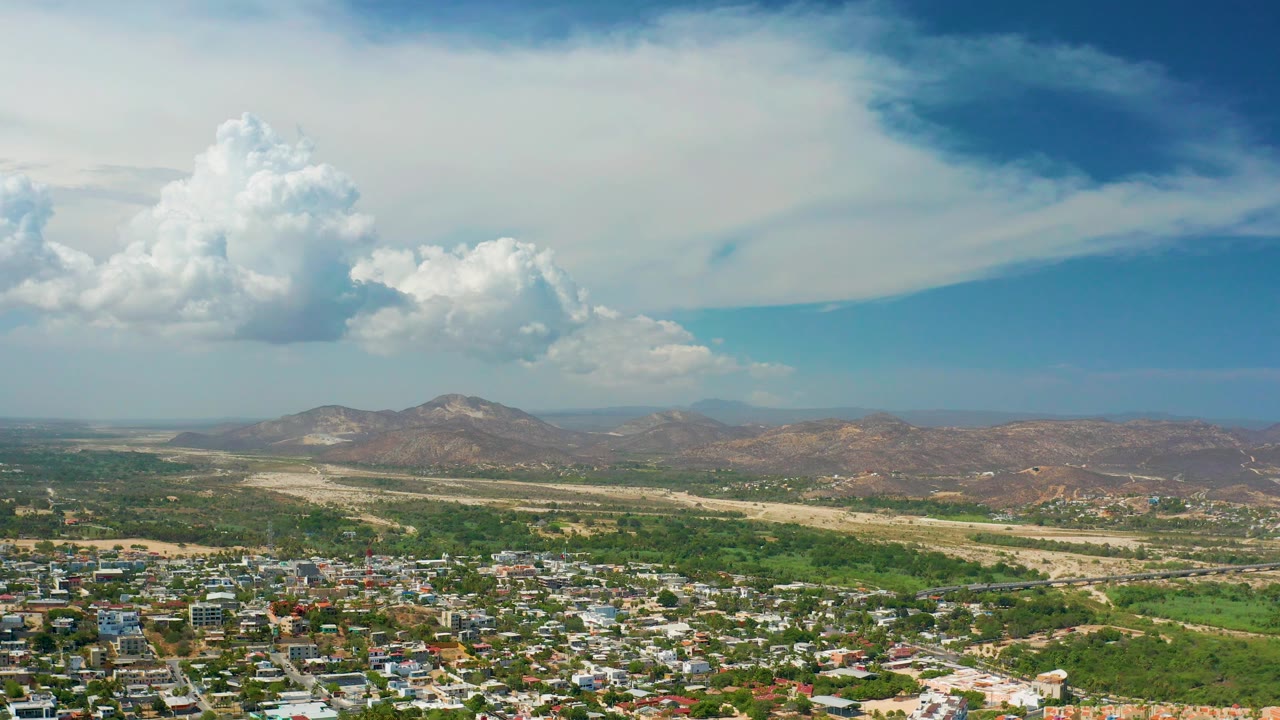 Opening aerial frame over Los Cabos, Mexico, with homes along the lower edge, green agricultural land, a pale dry wash, distant low mountains, large white clouds, and no visible people.