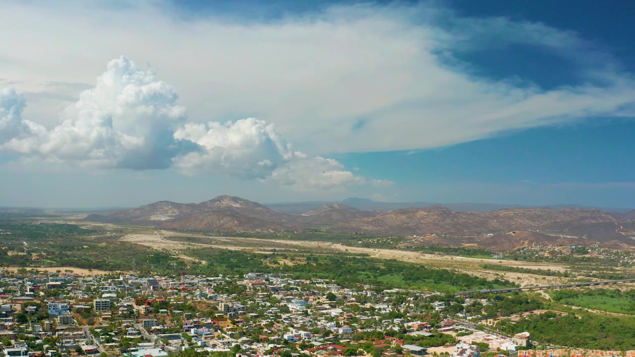 Wide Los Cabos aerial view with compact rooftops in the foreground, green fields across the valley floor, a light-colored dry wash, brown hills beyond, broad cloud cover, and no visible people.
