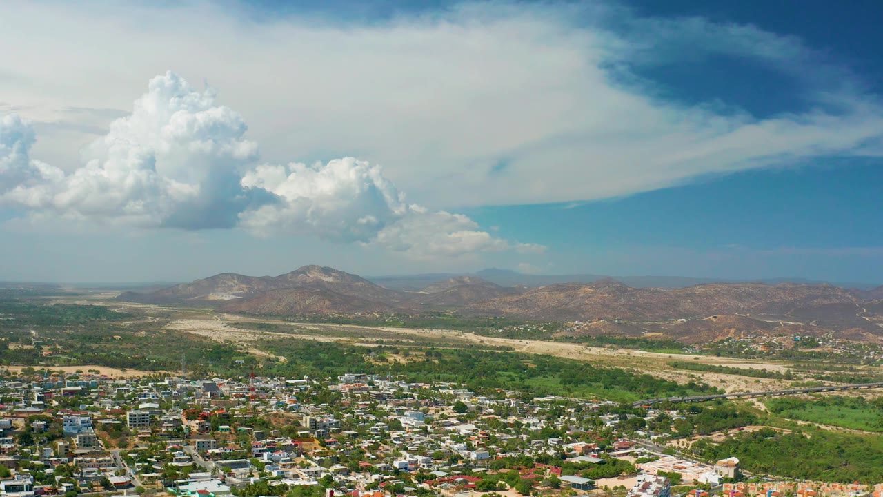 Los Cabos valley frame showing the town spread across the lower foreground, green cultivated areas, dry tan ground through the center, distant hills under a blue sky, and no visible people.
