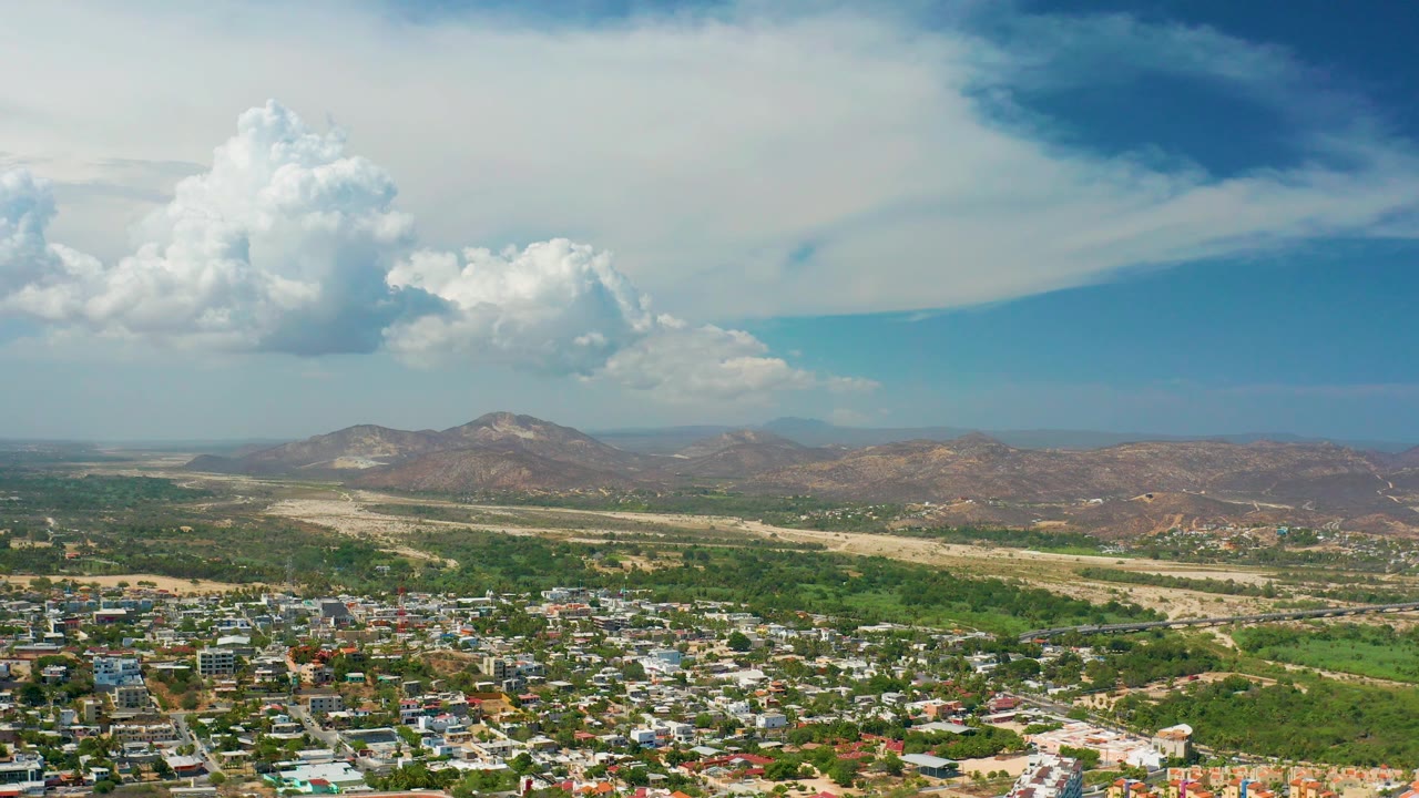 Town edge below green fields, pale dry wash, brown mountains, and bright clouds.