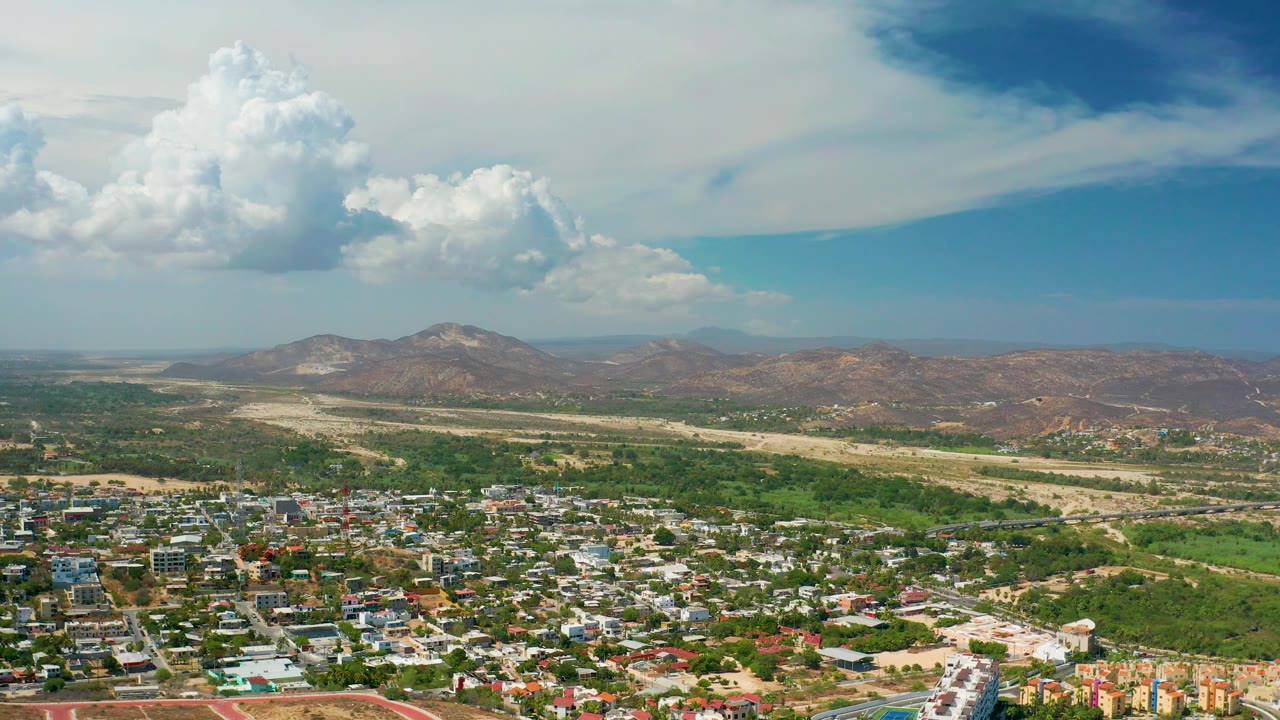 Wide aerial frame over Los Cabos, Mexico showing low-rise city blocks in the foreground, green vegetation beyond, dry open land, distant mountains, and no visible people.