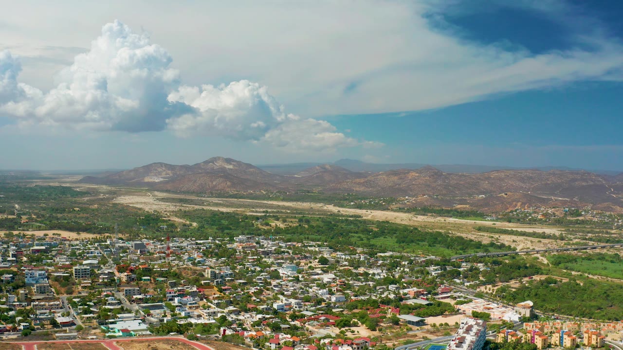 Aerial cityscape with rooftops, green corridor, dry terrain, mountains, and bright clouds.