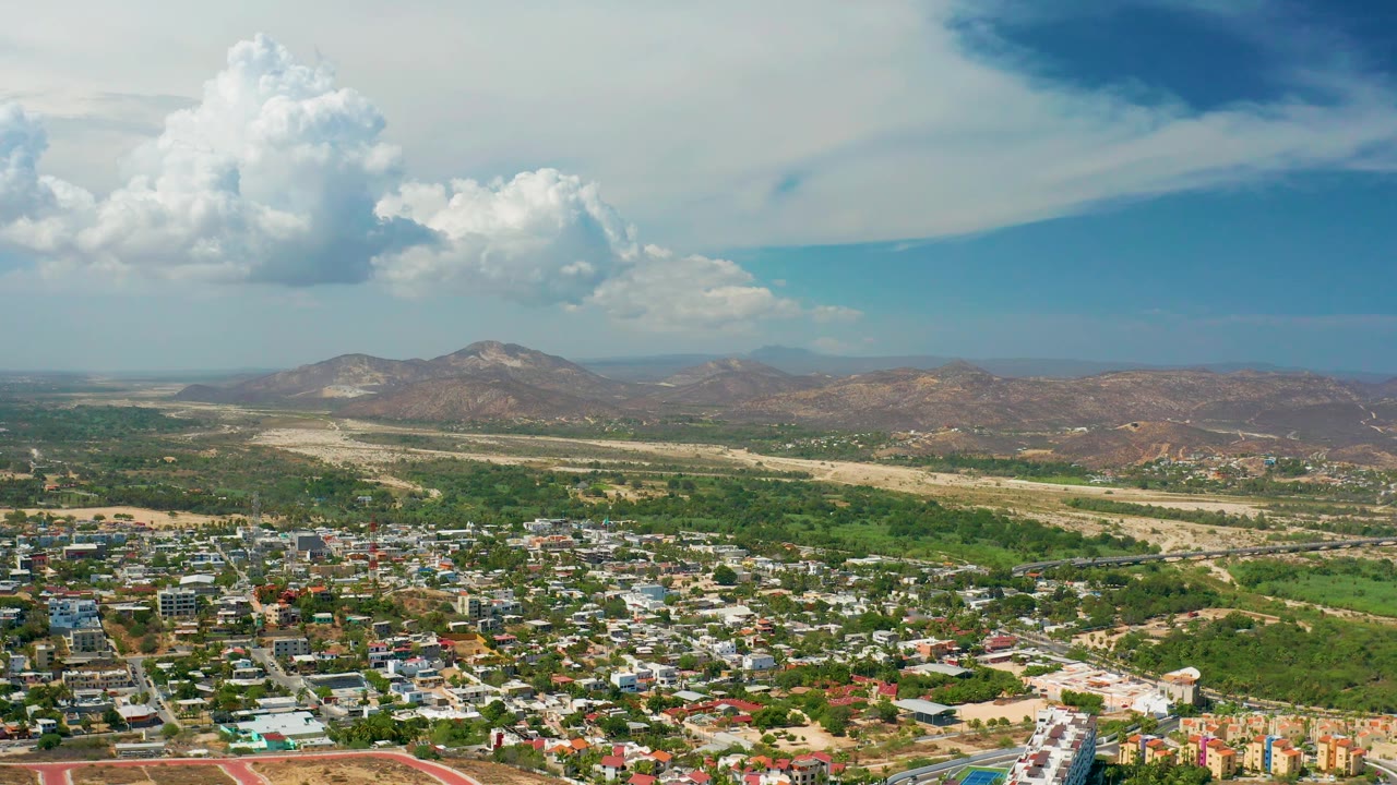 Los Cabos aerial frame showing a developed neighborhood below, broad open land beyond the buildings, low brown mountains in the background, soft daylight, and no visible people.