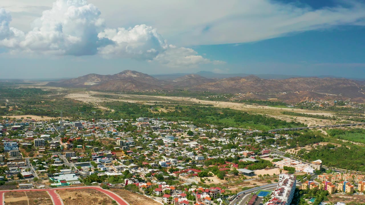 Los Cabos, Mexico aerial view showing a compact town in the foreground, patches of green vegetation, pale dry ground, rugged mountain ridges, blue sky, and no visible people.
