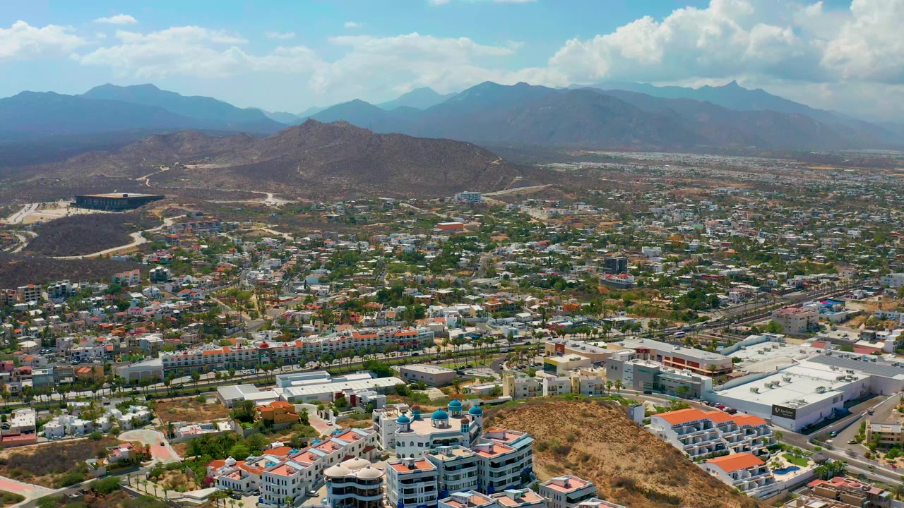 A high aerial frame over Los Cabos shows rooftops, roads, dry hills, distant mountains, bright clouds, and no visible people.