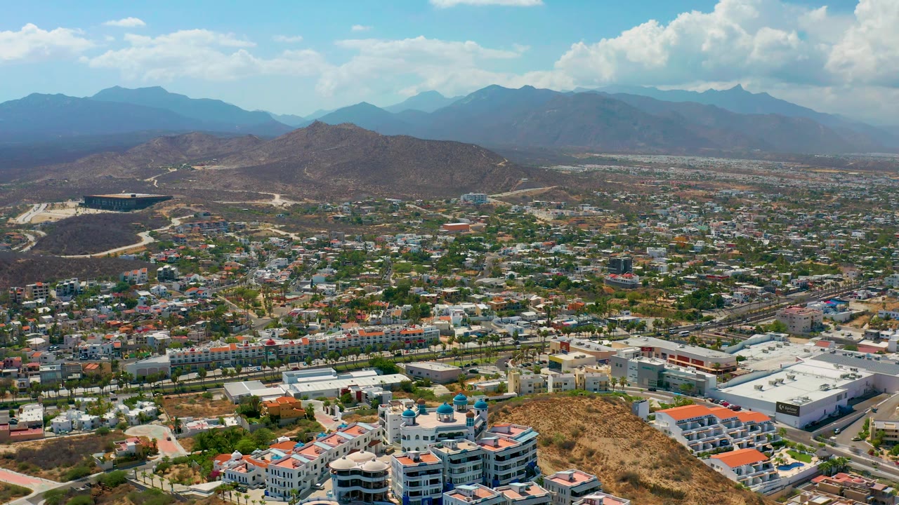 Aerial footage over Los Cabos shows foreground rooftops, a dense spread of buildings, dry ridges, soft blue mountains, and no visible people.