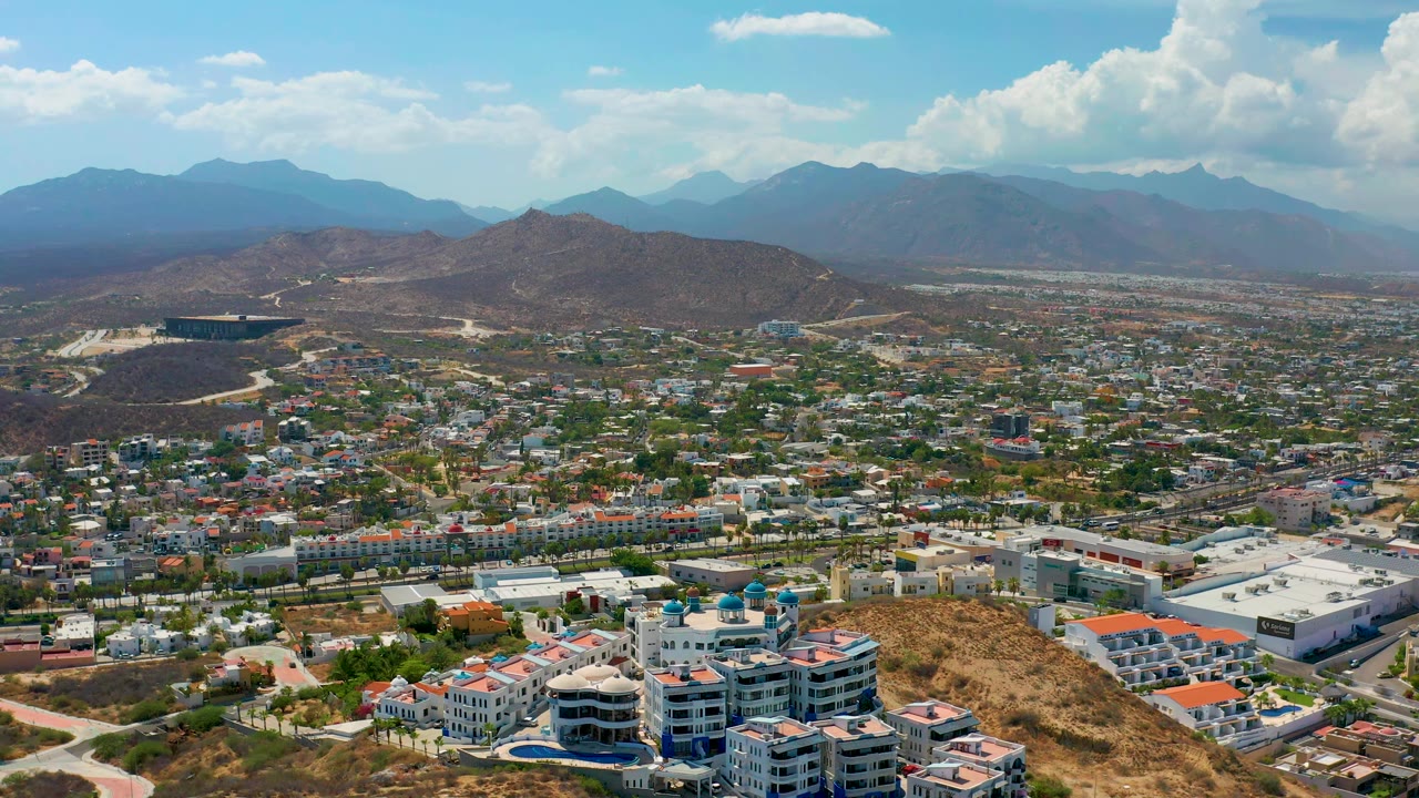 Aerial view over Los Cabos with hillside buildings in the foreground, a spread of roads and homes in the midground, hazy mountains beyond, and no visible people.