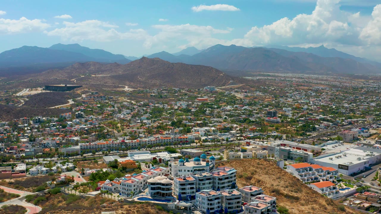 Los Cabos aerial frame showing white-roofed structures, residential-looking blocks, brown hills, a layered mountain range, bright clouds, and no visible people.