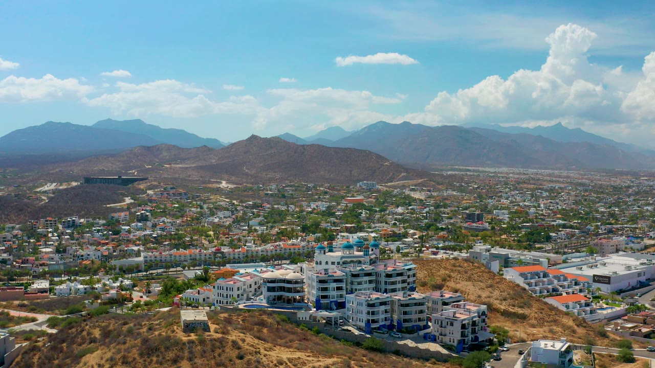 Wide aerial view over Los Cabos, Mexico, with white hillside buildings in the foreground, dense city blocks beyond, hazy mountains on the horizon, and no recognizable people.