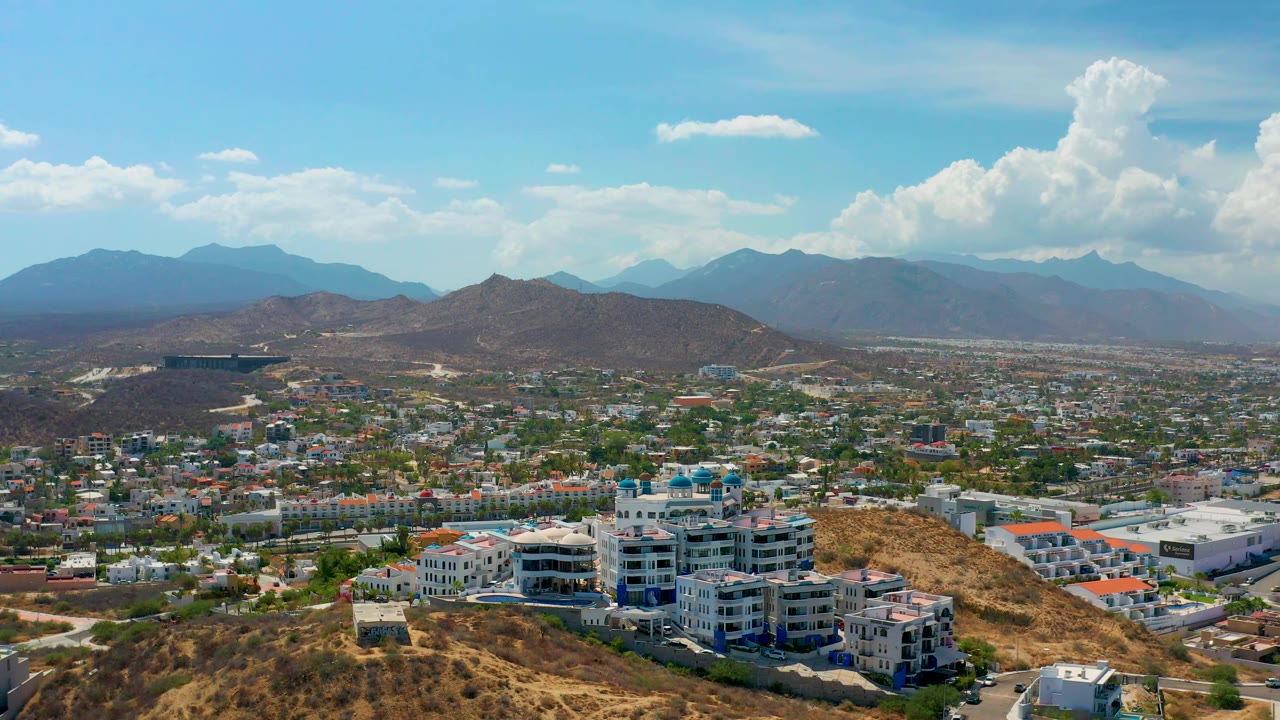 High aerial view over Los Cabos, Mexico, with a dry ridge in front, dense city streets and buildings across the valley, and mountains softened by haze in the background.