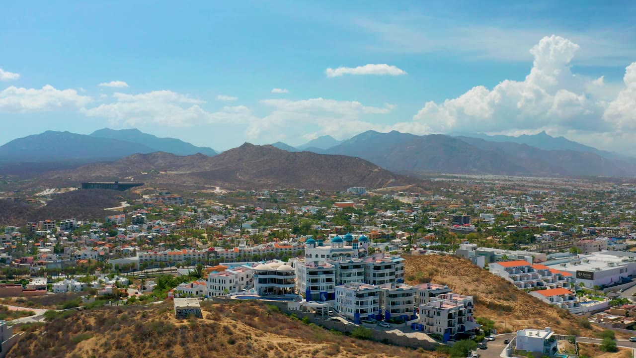 Wide aerial over urban Los Cabos with hills, roads, and clouds.