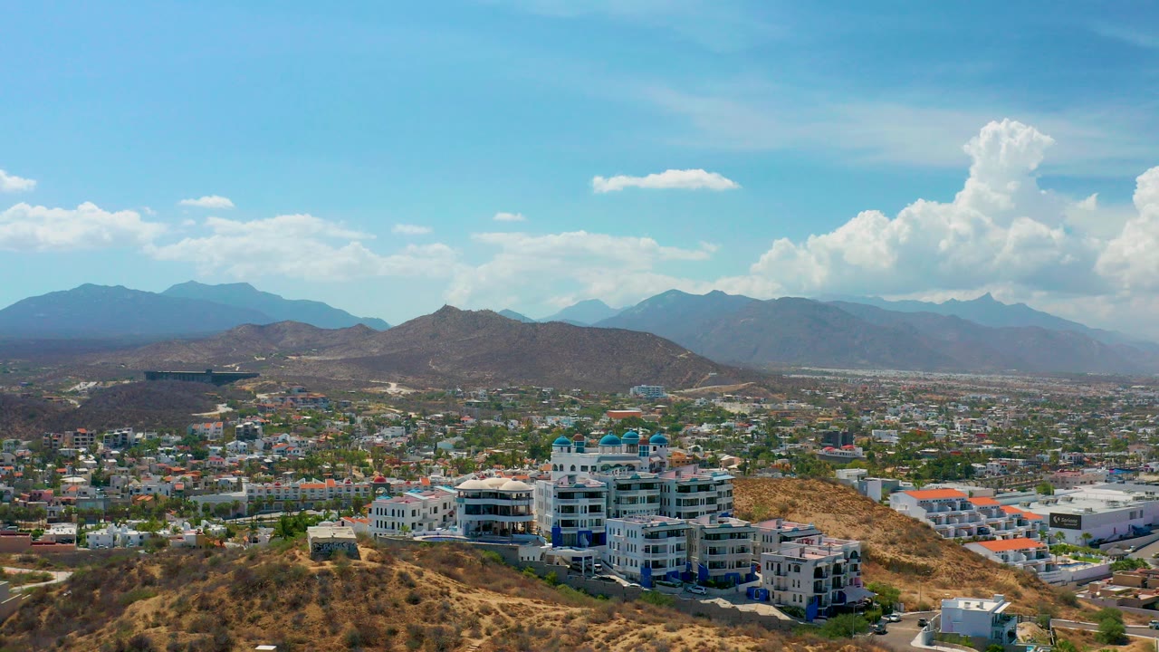 Opening aerial frame over Los Cabos with scrubby hills in front, white and gray building complexes below, distant city blocks, mountain ridges, bright clouds, and no visible people.