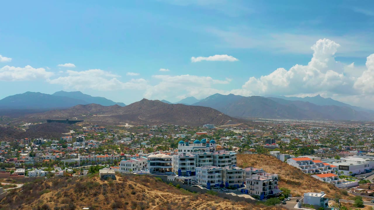 Wide Los Cabos aerial view with the building complex centered lower in frame, brown hills in the foreground, spread-out city development behind it, blue mountains, clouds, and no visible people.
