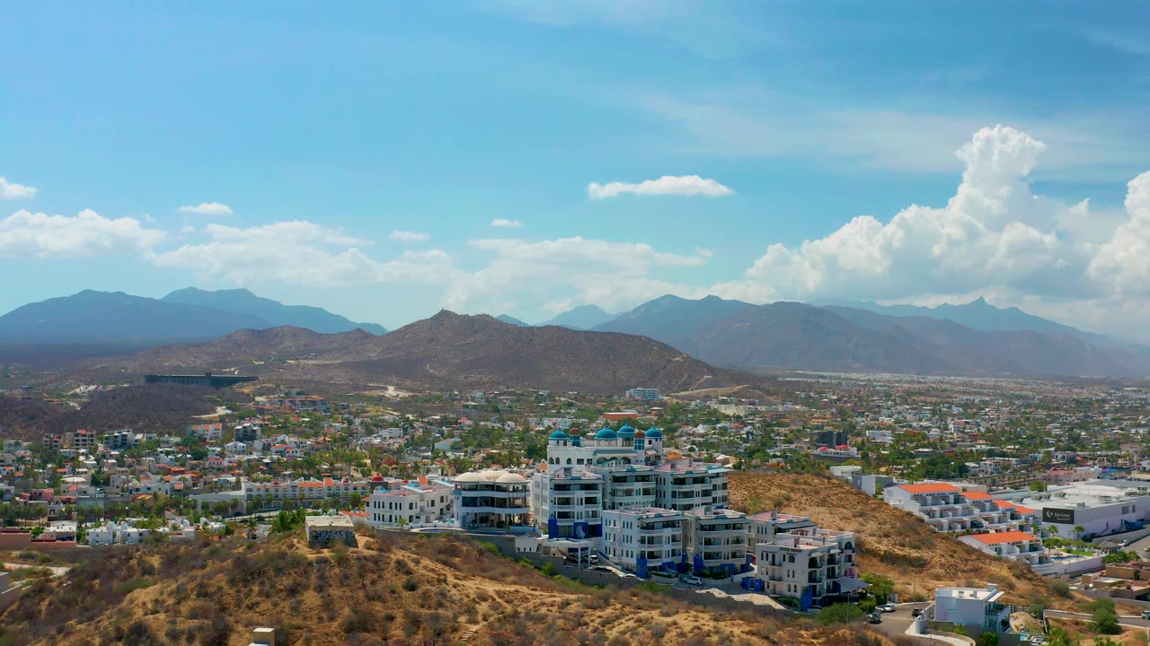 Los Cabos aerial frame showing white hillside buildings near the lower center, dry terrain around them, a dense low city grid, layered mountain ridges, high white clouds, and no visible people.