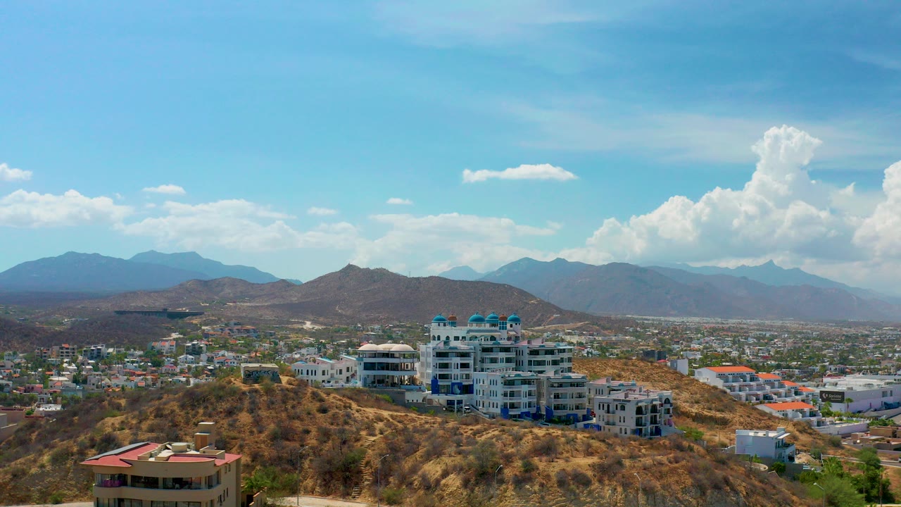 Los Cabos hillside view with white and blue multi-story buildings, dry terrain, distant mountains, bright clouds, and no visible people.