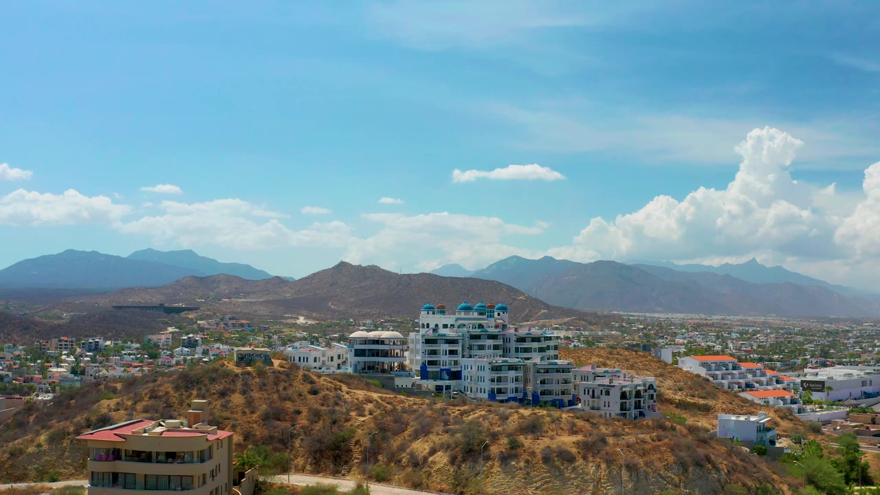 Los Cabos view across brown hills and pale city buildings, with a prominent modern complex, distant mountain layers, and no visible people.