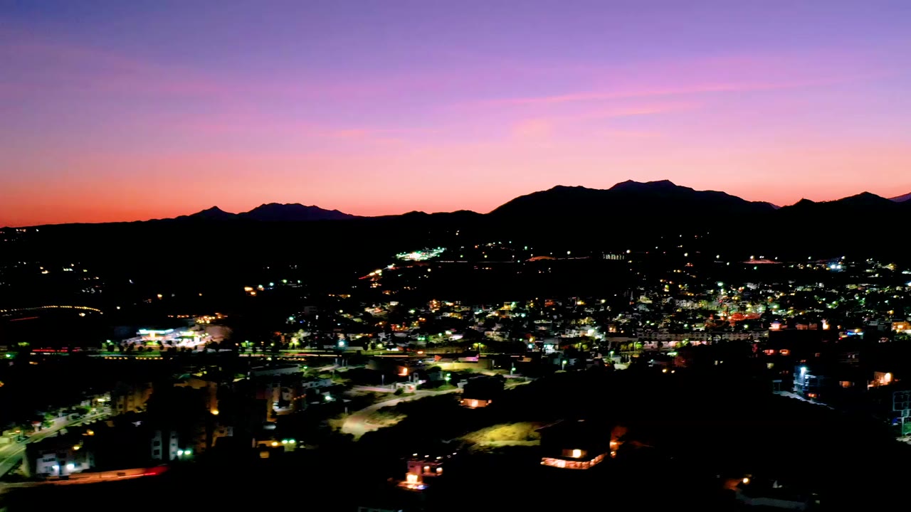 Elevated city lights below a purple sunset sky and dark mountain ridge.