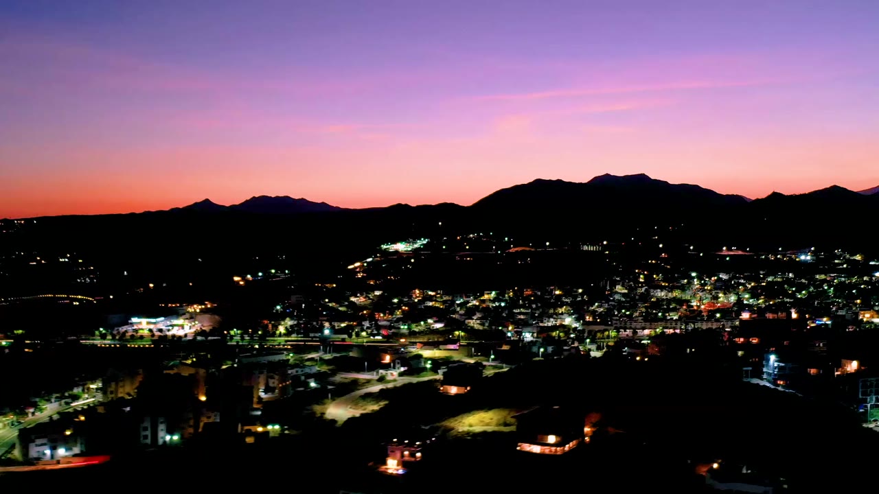 Los Cabos, Mexico appears from an elevated view with dense city lights, a black mountain ridge, and a warmer orange band along the horizon, with no visible people.