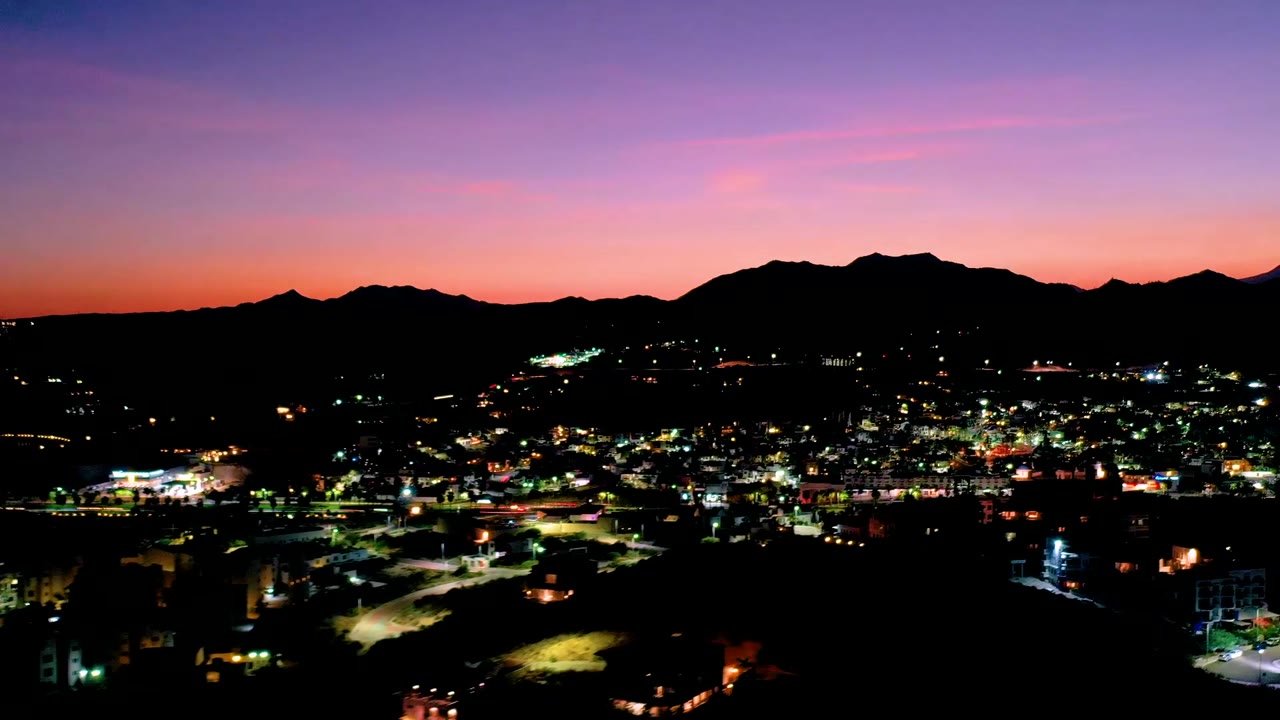 A wide Los Cabos, Mexico dusk view shows scattered bright city lights beneath a long silhouetted mountain range and pink-purple sky, with no visible people.