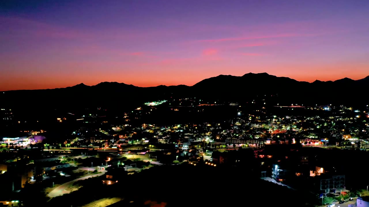 Los Cabos, Mexico city blocks and roads glow below a dark mountain horizon as the sky shifts from orange near the ridge to violet above, with no visible people.