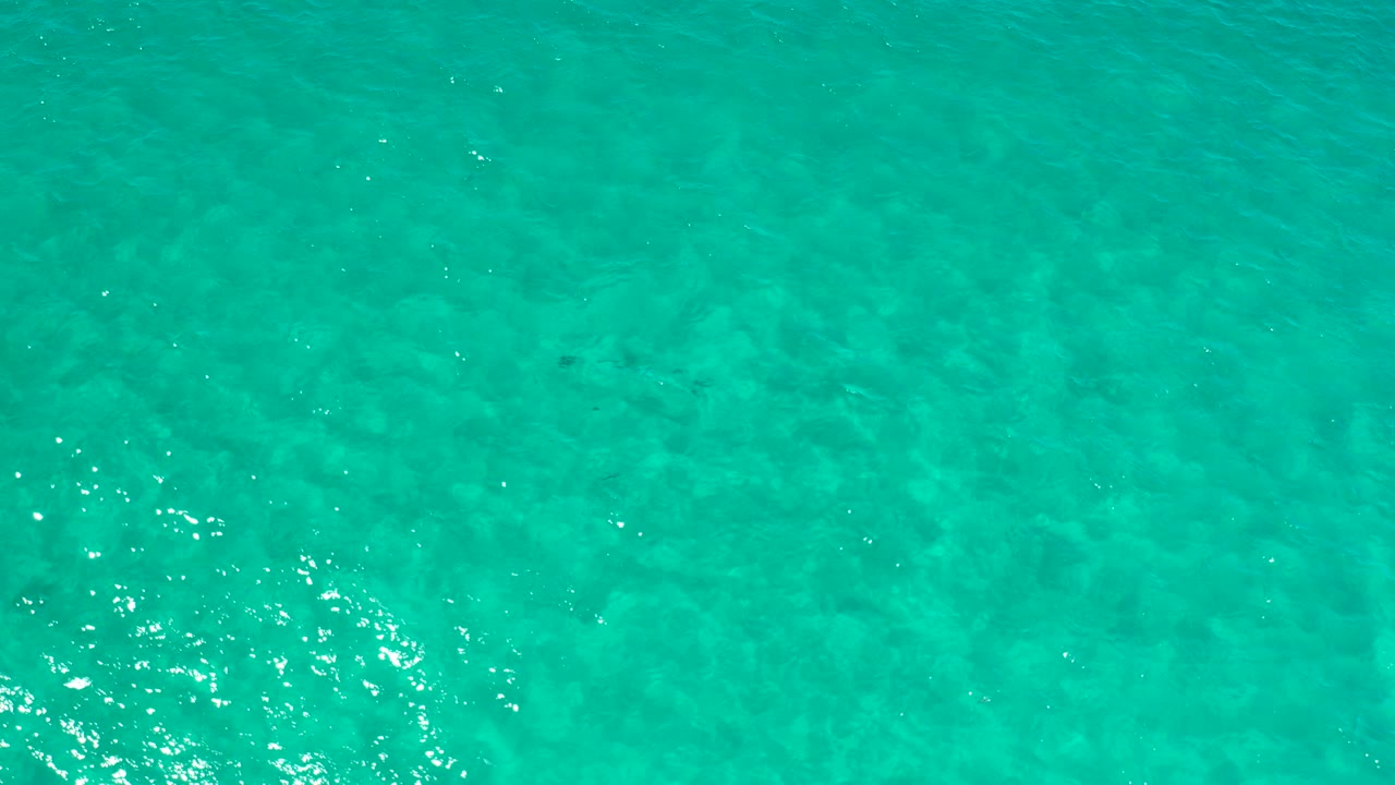 Overhead view of turquoise ocean water in Los Cabos, Mexico, with faint seabed texture, scattered sunlight glints, and no visible people.