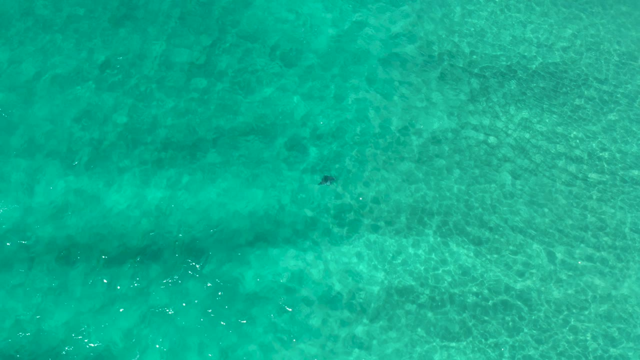 Clear blue-green water in Los Cabos shows faint wave lines, mottled underwater shadows, bright surface glints, and no visible people.