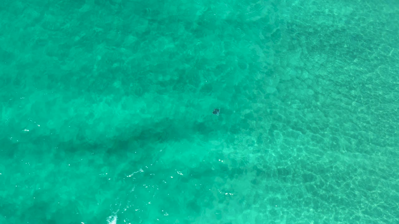 The overhead frame shows Los Cabos turquoise water crossed by darker underwater bands, tiny white wave flecks, and no visible people.