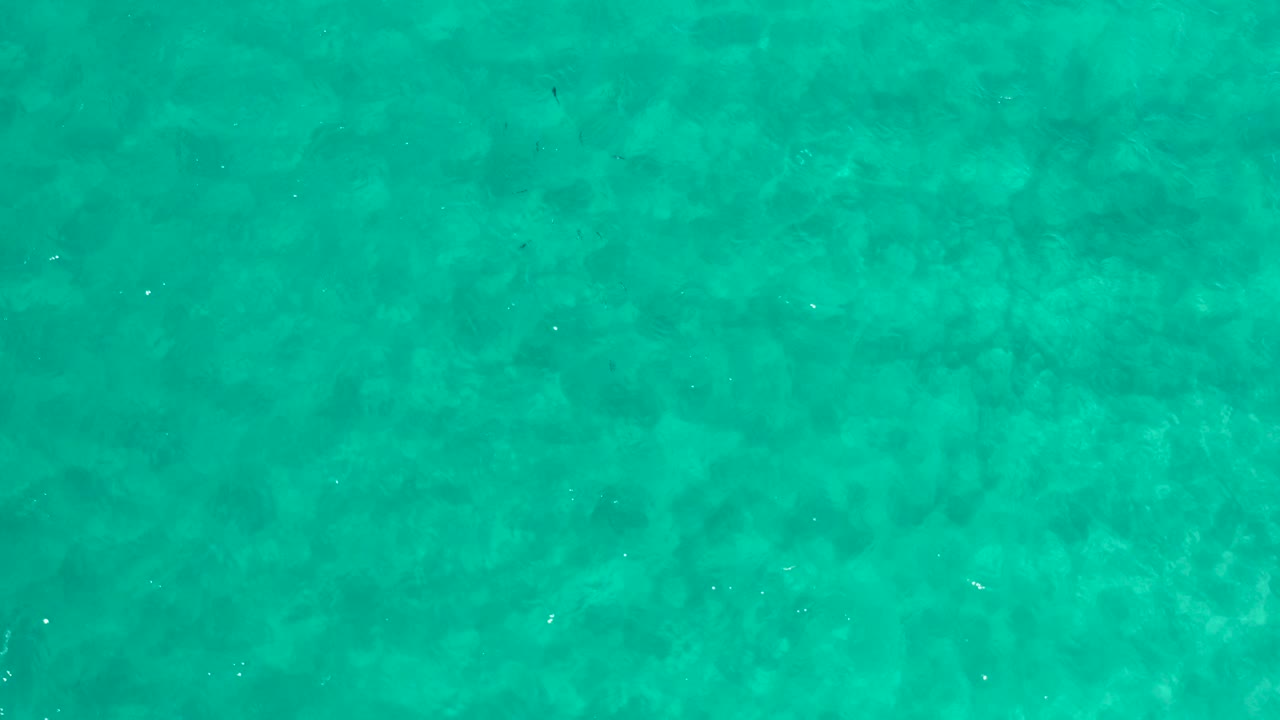Overhead view of clear turquoise ocean water in Los Cabos, Mexico, with faint darker patches below the surface and no visible people.