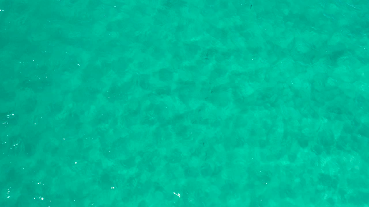 Top-down view of turquoise water in Los Cabos with faint submerged darker patches, small surface glints, and no visible people.