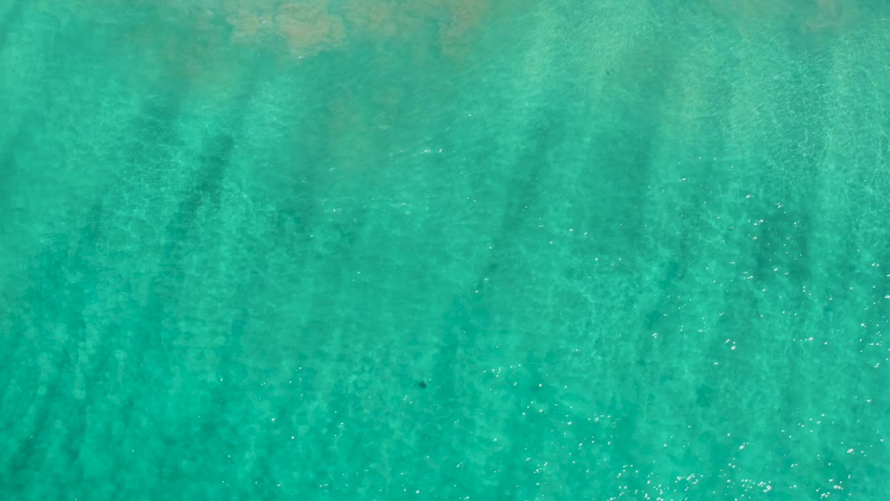 Clear green-blue sea fills the frame in a top-down Los Cabos view, with pale sand beneath the ripples and no visible people.
