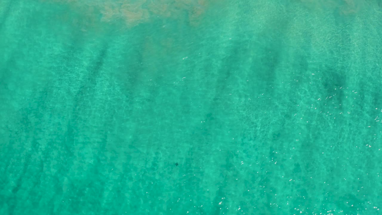 Aerial view of shallow turquoise ocean in Los Cabos, Mexico, showing pale sandy bottom, soft wave texture, and no visible people.