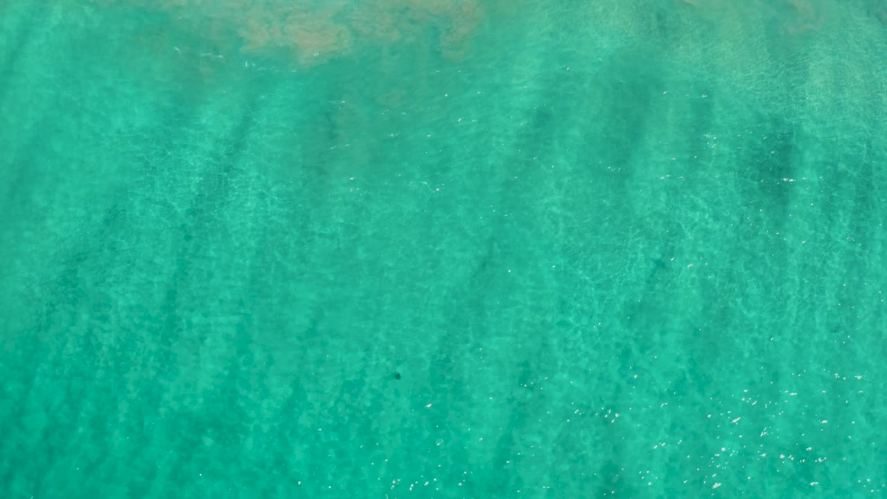 Top-down frame of clear Los Cabos water with faint darker underwater marks across the sandy bottom and no visible people.