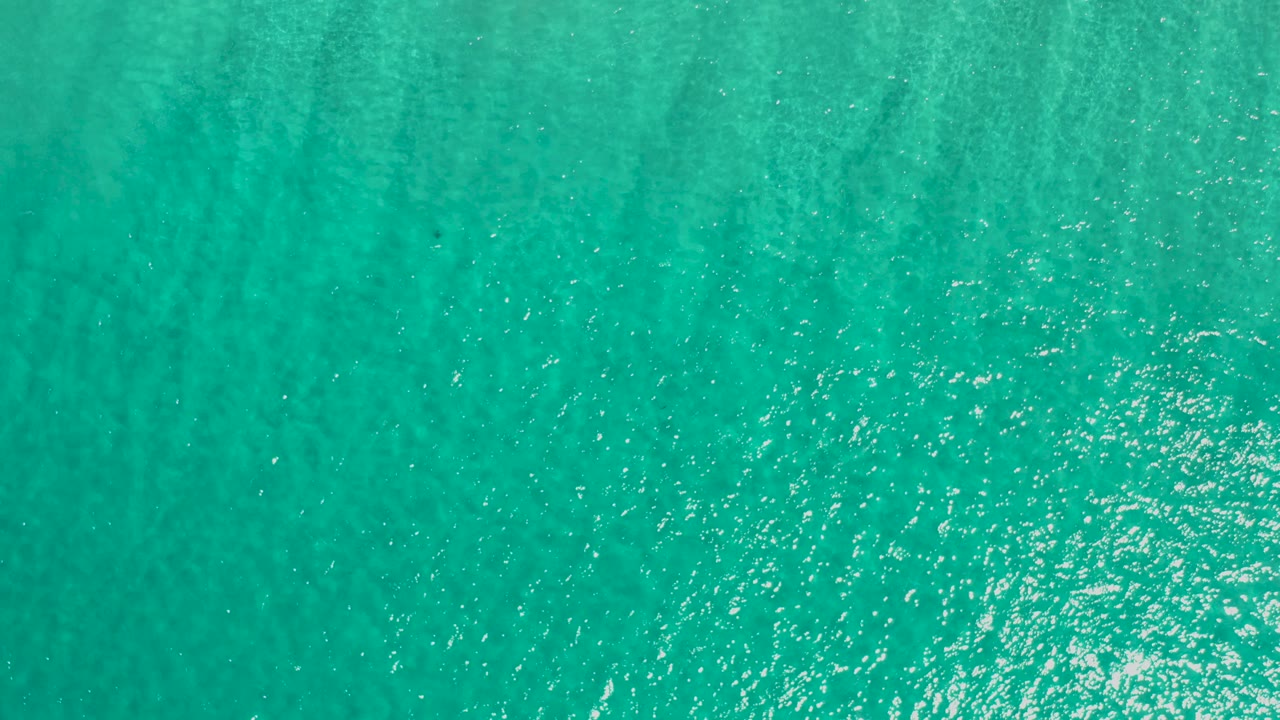 Top-down view of turquoise ocean water in Los Cabos, Mexico, with fine ripple texture, small bright reflections, and no visible people.