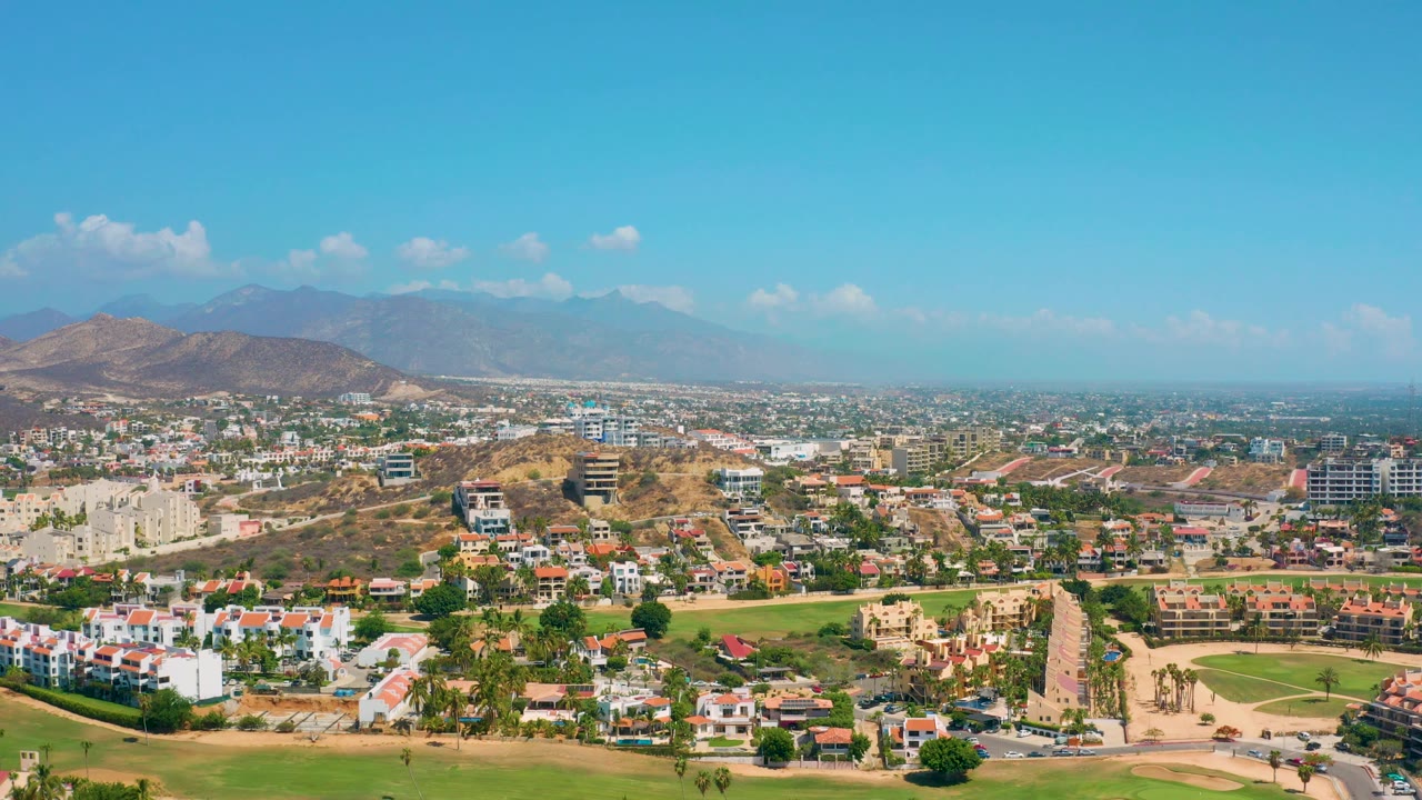 High view across Los Cabos homes, green patches, tan roads, arid slopes, a hazy mountain horizon, scattered clouds, and no visible people.