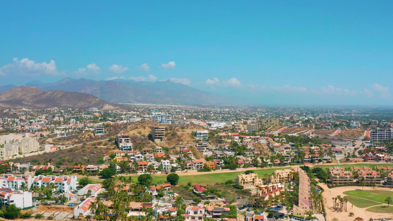 Opening aerial frame over Los Cabos, Mexico, showing a broad suburban area, green lawns, dry hills, distant mountains, blue sky, and no visible people.