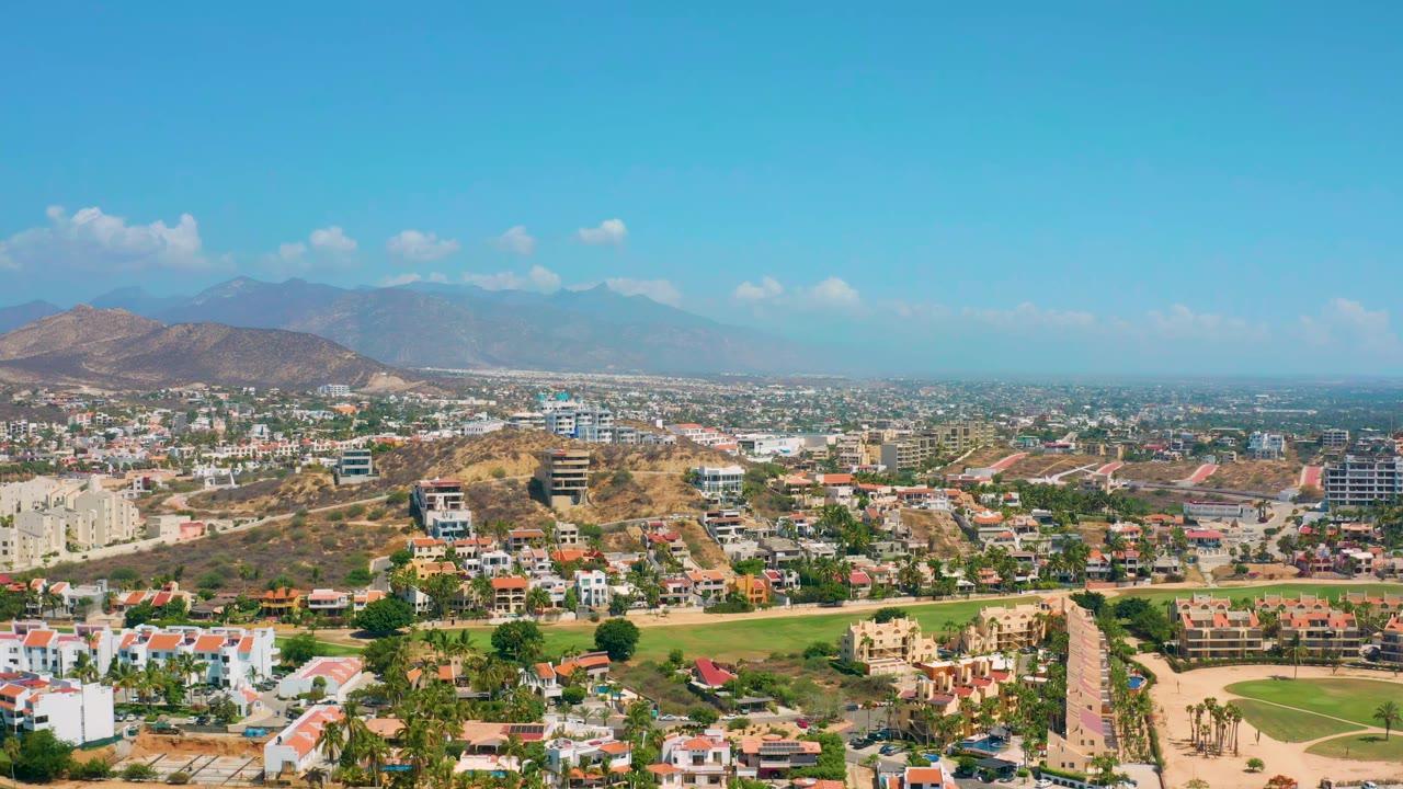 Second frame over Los Cabos, Mexico, with clustered houses, a straight road through the foreground, dry hills, distant mountains, bright sky, and no visible people.