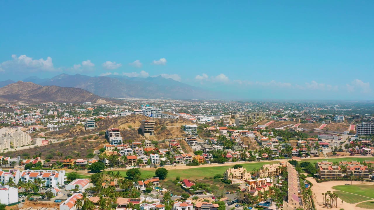 Broad aerial frame of a sunny suburb with roads, homes, hills, and mountains.