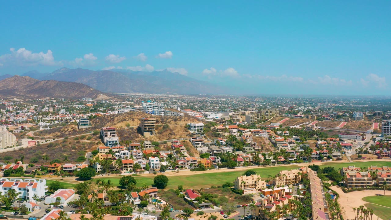 Opening aerial frame over Los Cabos with upscale homes, tan hills, a green landscaped strip, distant mountains, blue sky, and no visible people.