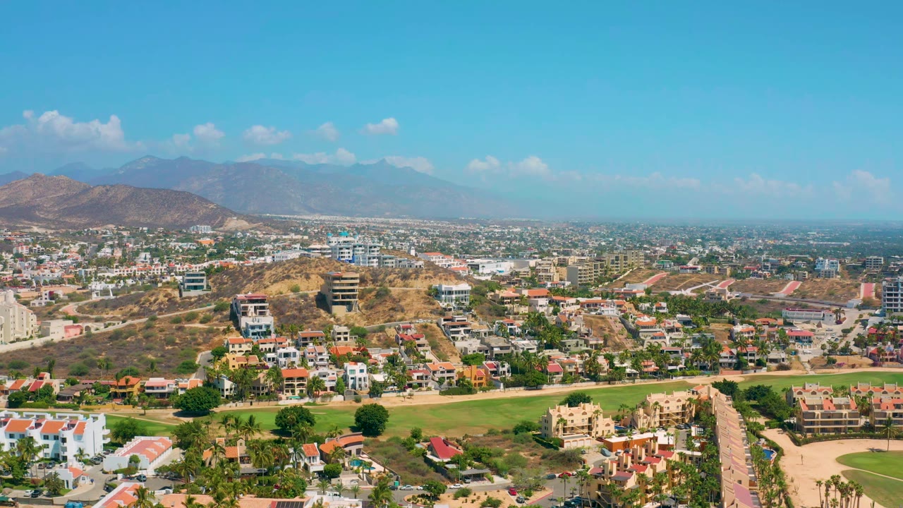 Early aerial view in Los Cabos showing hillside residences, low-rise buildings, green fairway-like landscaping, hazy mountains, and no visible people.
