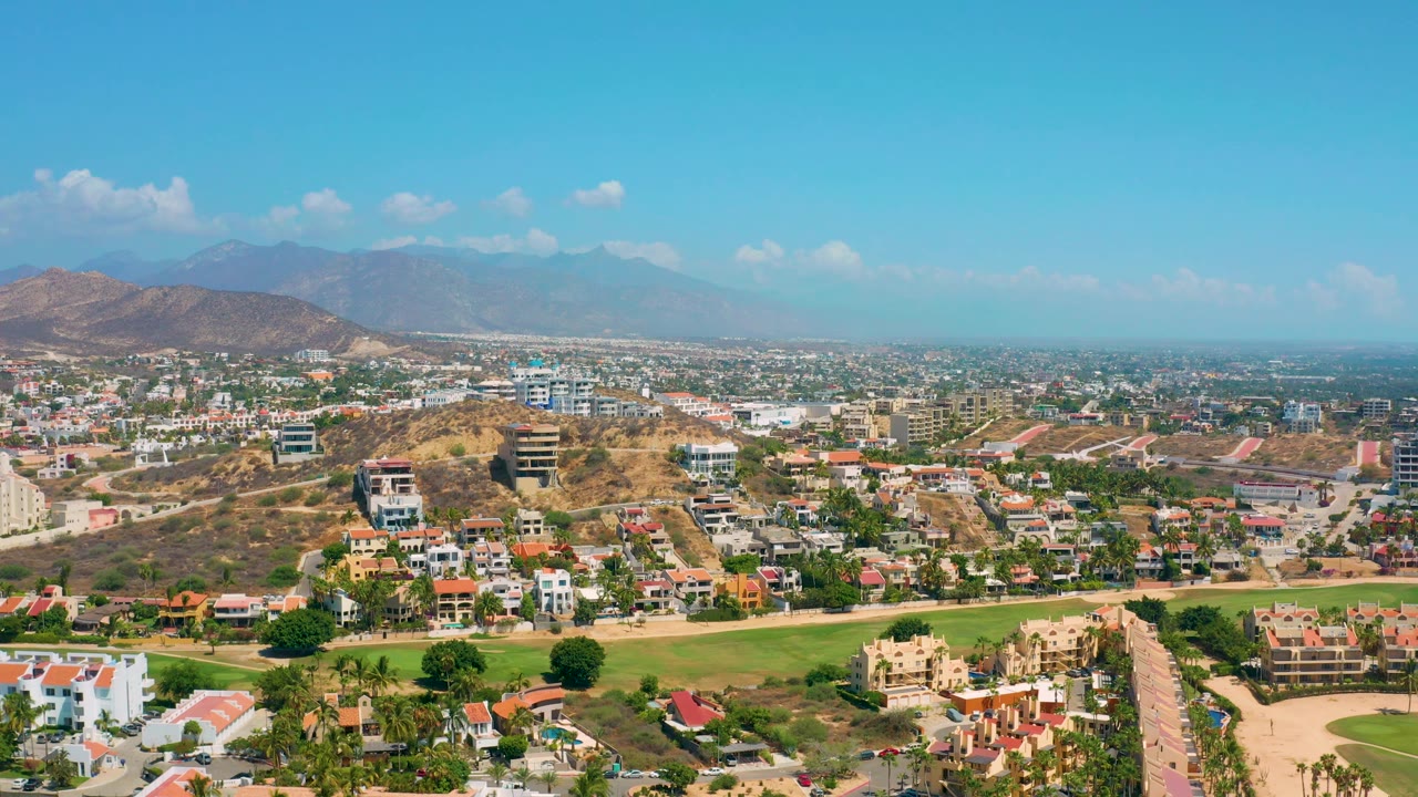 Los Cabos aerial frame with clustered homes and roads in front, dry slopes through the center, a mountain range on the horizon, and no visible people.