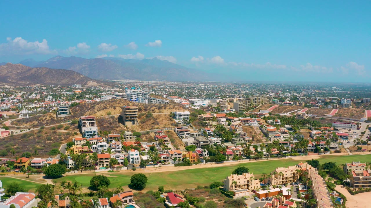 Opening frame of Los Cabos, Mexico from above, showing hillside houses, green fairway grass, distant town blocks, hazy mountains, scattered clouds, and no visible people.