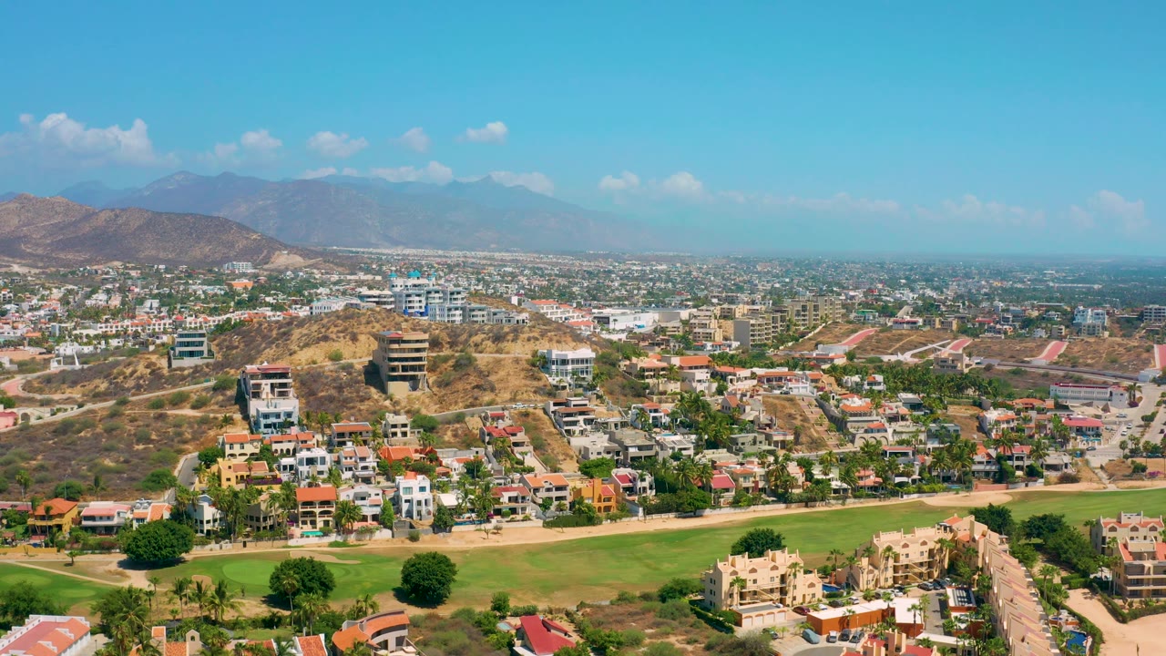 Los Cabos, Mexico aerial still showing a residential ridge above green grass, denser buildings across the plain, mountain silhouettes in haze, and no visible people.