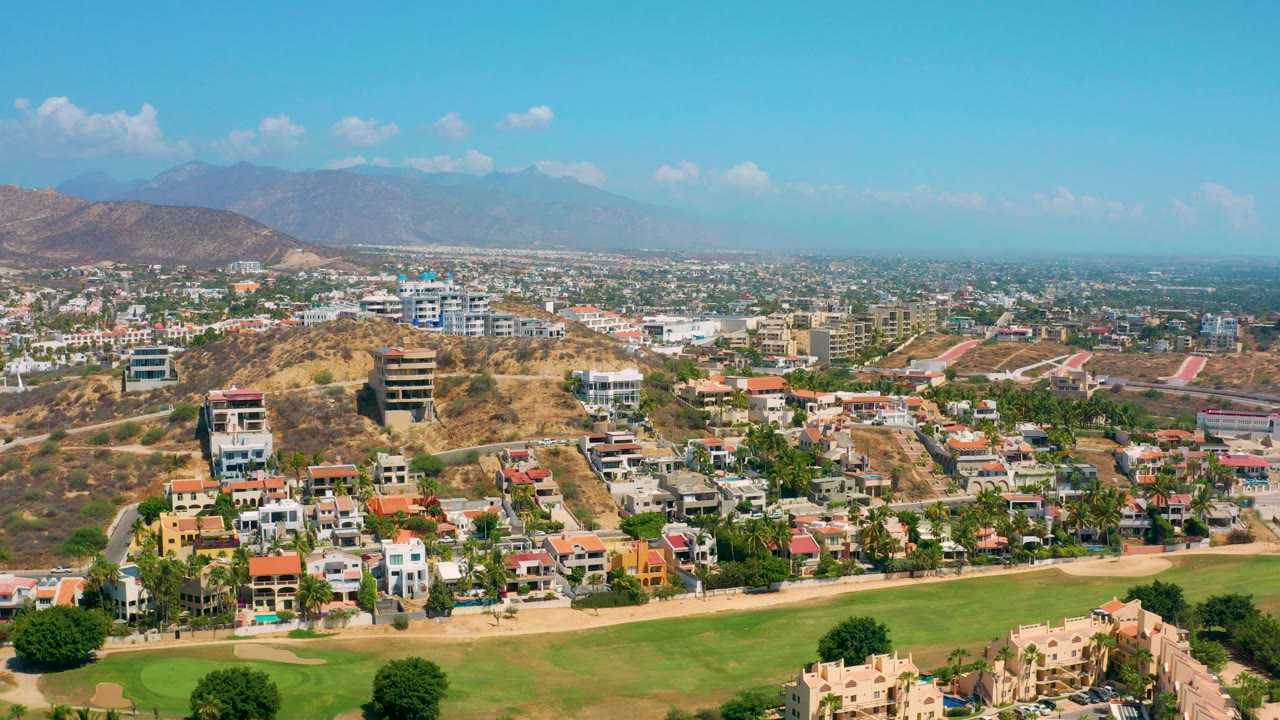 Wide aerial view over Los Cabos hillside homes with green golf-course turf at the bottom, low-rise buildings across the valley, hazy mountains in back, and no visible people.